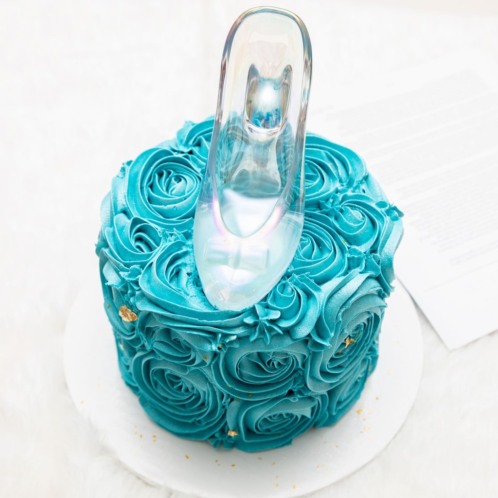 Photo of a blue cake with gold decorations on top of which is a glass slipper, all set upon a white cloth.  