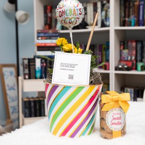 Image of a rainbow bucket with yellow roses, a congrats balloon and a sweet smiles jar of cookies.
