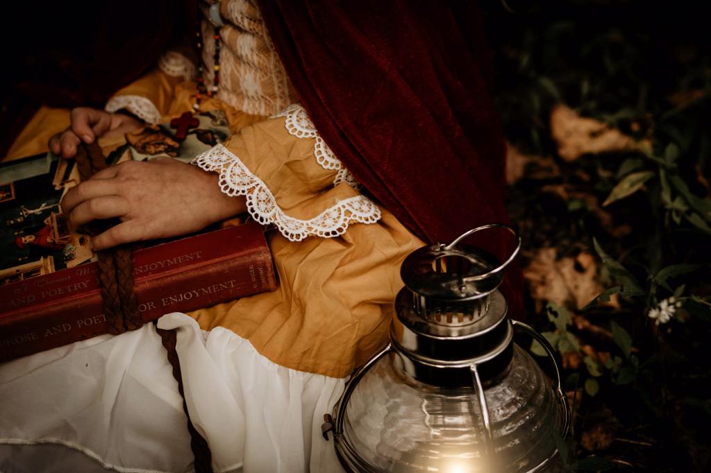 Photo of the midsection of a child wearing a yellow dress with a red velvet cape holding a stack of books with a lantern nearby.
