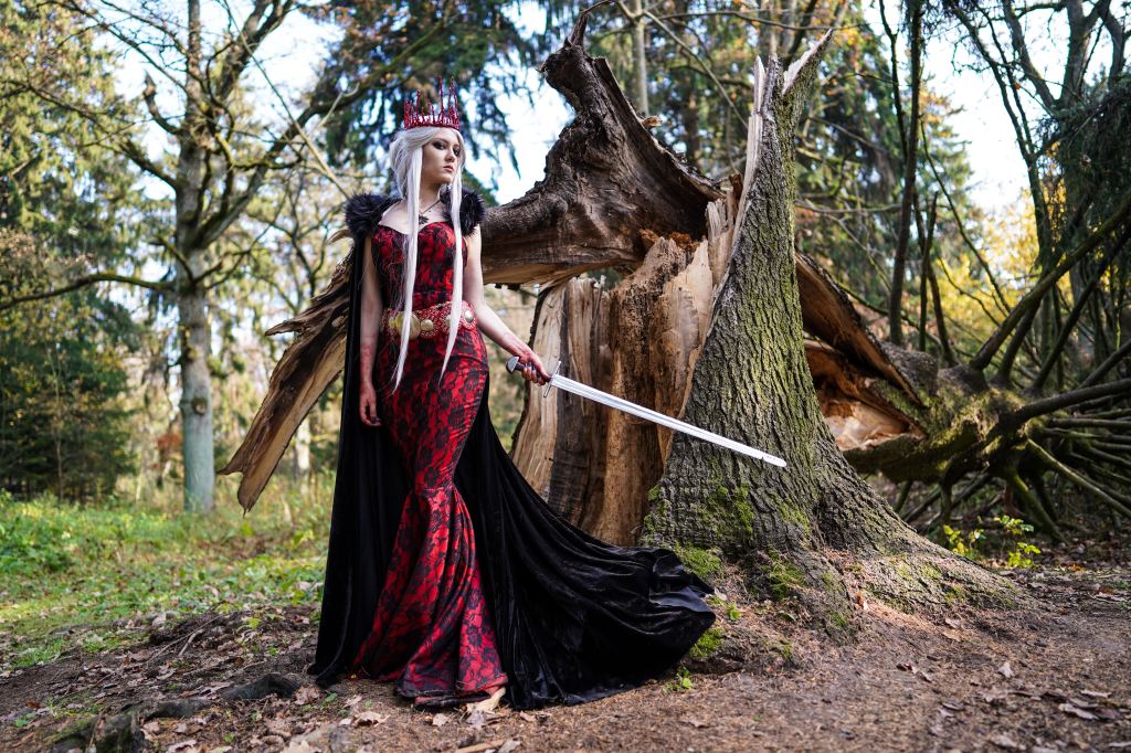 Photo of a white woman with silver blond hair wearing a red crown, a red and black gown with a long black cape, wielding a sword in front of a fallen tree.