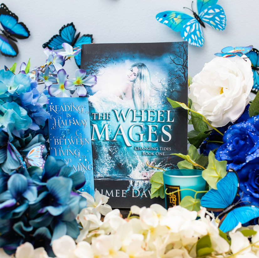 Photo of The Wheel Mages surrounded by fake white and blue flowers and blue butterflies along with a blue bookmark that says Reading is Halfway Between Living and Dreaming. 
Copyright: Aimee Davis
Source: Instagram @writingwaimee 