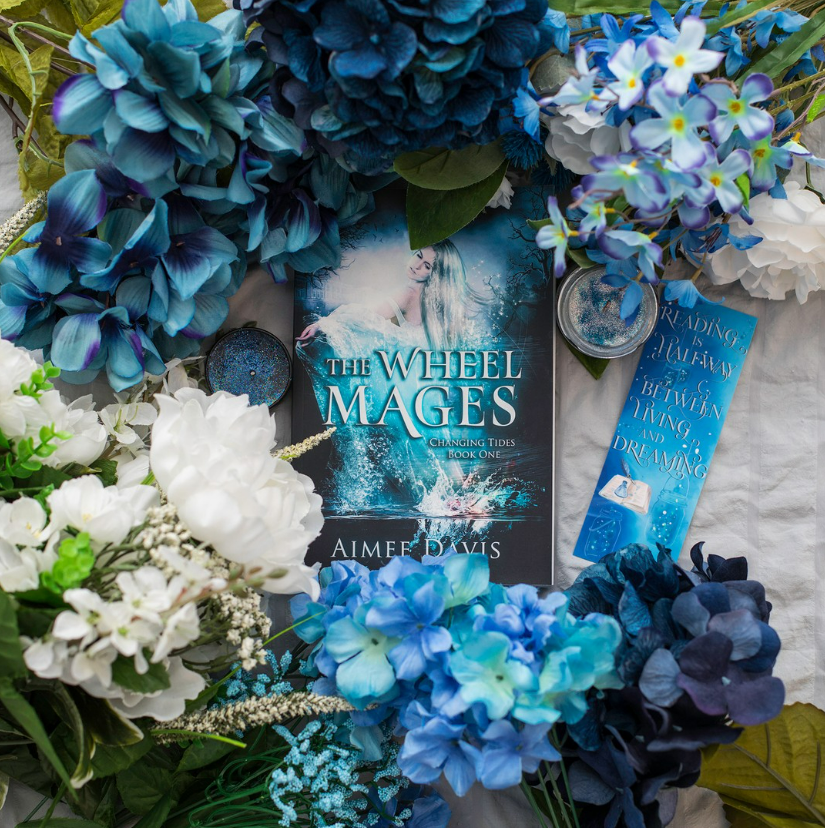 Photo of The Wheel Mages lying flat with white and blue flowers around it and blue sparkly candles along with a bookmark that says reading is halfway between living and dreaming. 
Copyright: Aimee Davis
Source: Instagram @writingwaimee