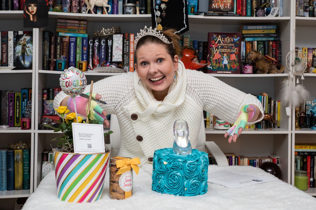 Photo of a white woman (me) in a white sweater wearing rainbow gloves and a crown gesturing toward a contract and a blue cake with a Cinderella slipper on it. 
Copyright: Aimee Davis 