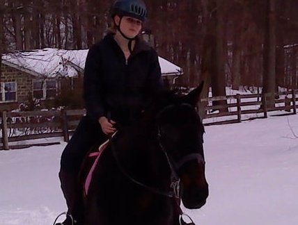 A woman (me) with a green riding helmet rides a bay horse with a pink saddle pad in the snow.