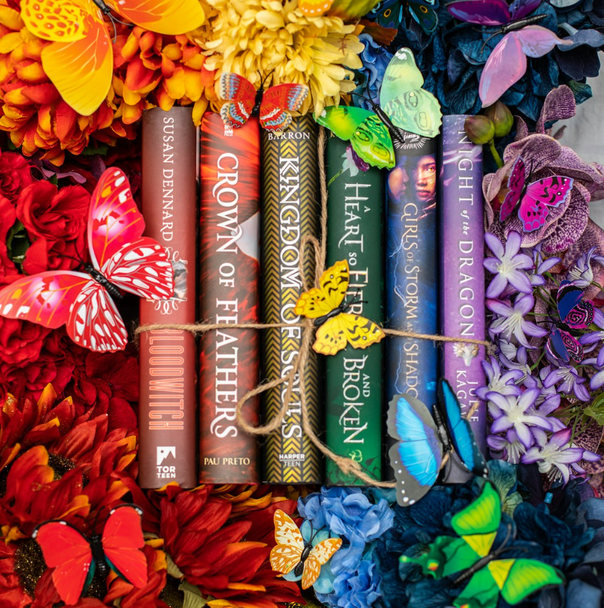 Photo of a rainbow of book spines including Bloodwitch, Crown of Feathers, Kingdom of Souls, A Heart so Fierce and Broken, Girls of Storm & Shadow, and the Night of the Dragon. All surrounded by rainbow flowers and butterflies.
(c) Aimee Davis @writingwaimee