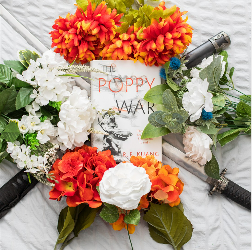 A photo of The Poppy War by R. F. Kuang lying atop two Katanas surrounded by fake orange and white flowers. 
© Aimee Davis (@writingwaimee on Instagram) 