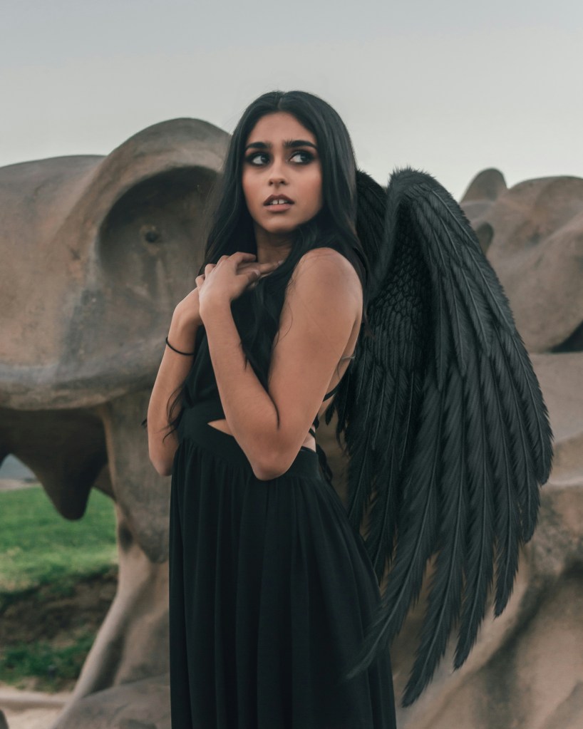 Photo of a brown girl with long black hair in a black gown sporting black wings standing in front of a dragon statue, hands pressed to chest, looking into the distance.
© Andre Sebastian, Unsplash