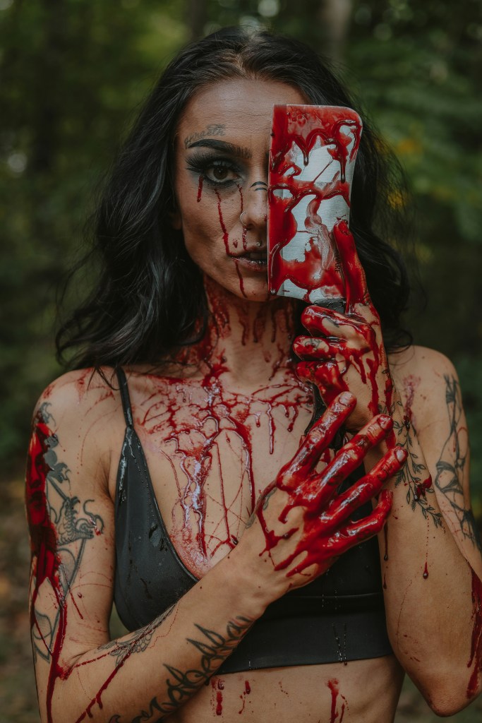 Photo of a brown-skinned woman with long black hair holding a butcher's knife over one eye, covered in blood. © Sierra Koder, Unsplash